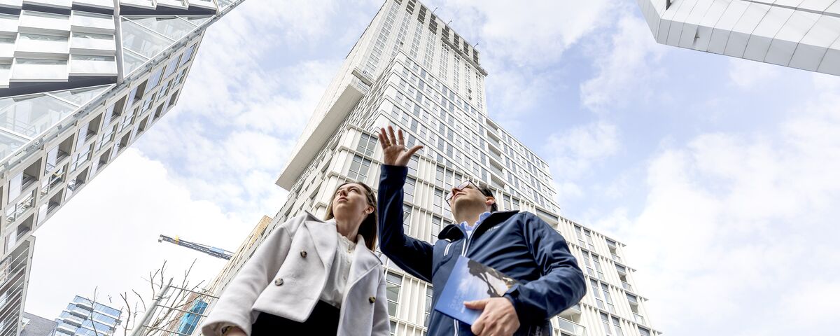 Man and woman in front of grey high-rise building looking up and pointing.